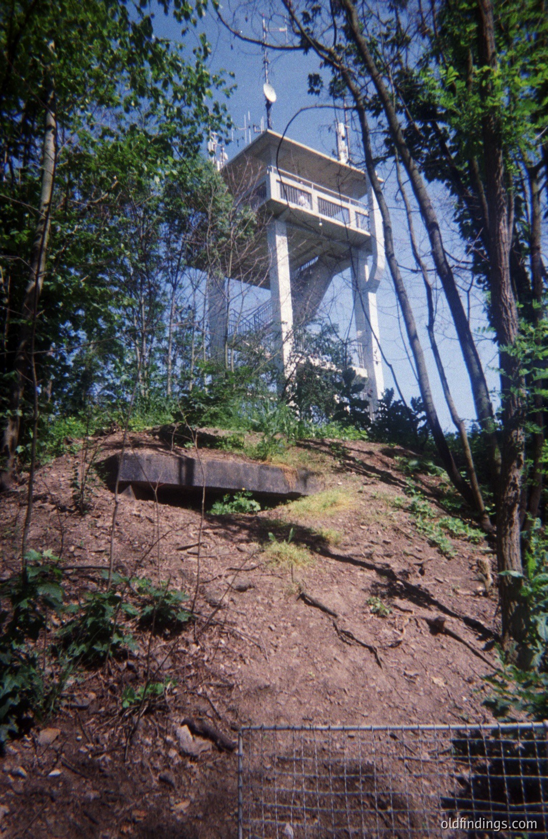 A tall, concrete observation tower rises above a wooded hillside. The tower features a circular platform and multiple antennas. A concrete structure sits at the base of the hill, partially obscured by vegetation and a wire fence. Likely mid-20th century construction. Possible military or communications site.