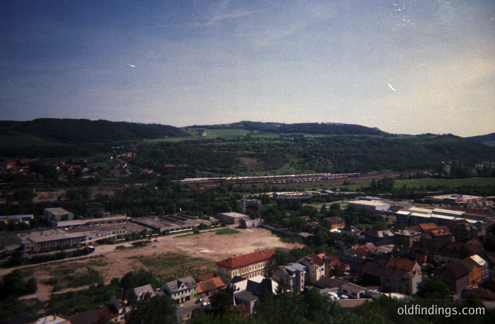 Elevated view of a town featuring red-roofed buildings, a rail yard, and a wooded hillside landscape. Likely a post-industrial scene with a mix of residential and commercial structures. The color palette suggests a mid-20th century film photograph. Appears to be a European location.