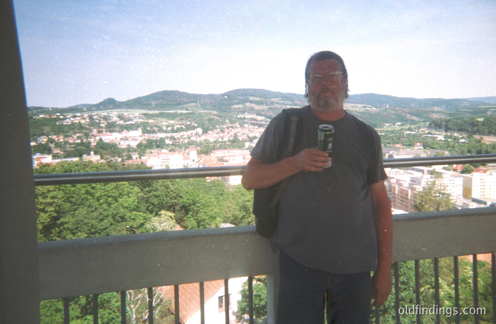 A man stands on a balcony overlooking a town and forested hills. He wears a dark t-shirt and jeans, holding a beverage. The town features red-tiled roofs and apartment blocks. Likely tourist snapshot; circa 1990s. Geographic location uncertain. Potential stock image for travel/lifestyle content.