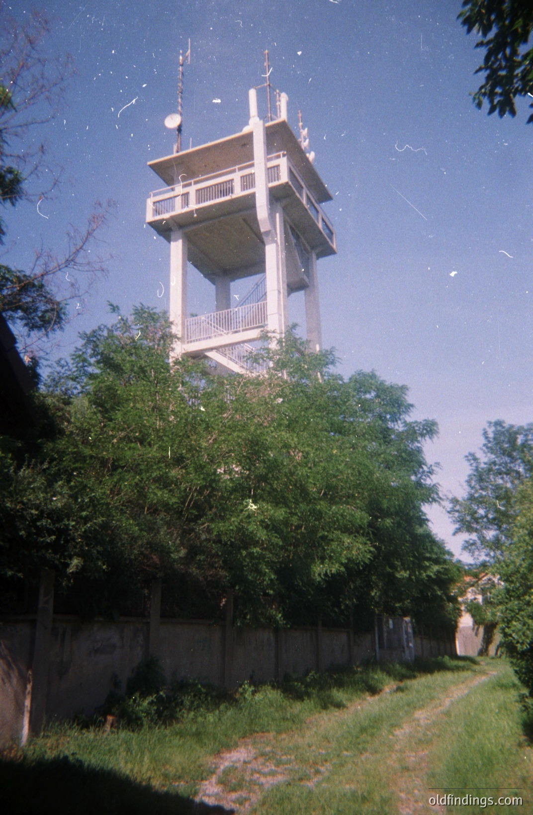 A tall, concrete observation tower rises above a fence and lush foliage. Likely built during the communist era, it features multiple levels with railings and antennas. A dirt path leads towards the tower, suggesting public access. The style points to 1970s or 80s architecture. Possible location: Eastern Europe.