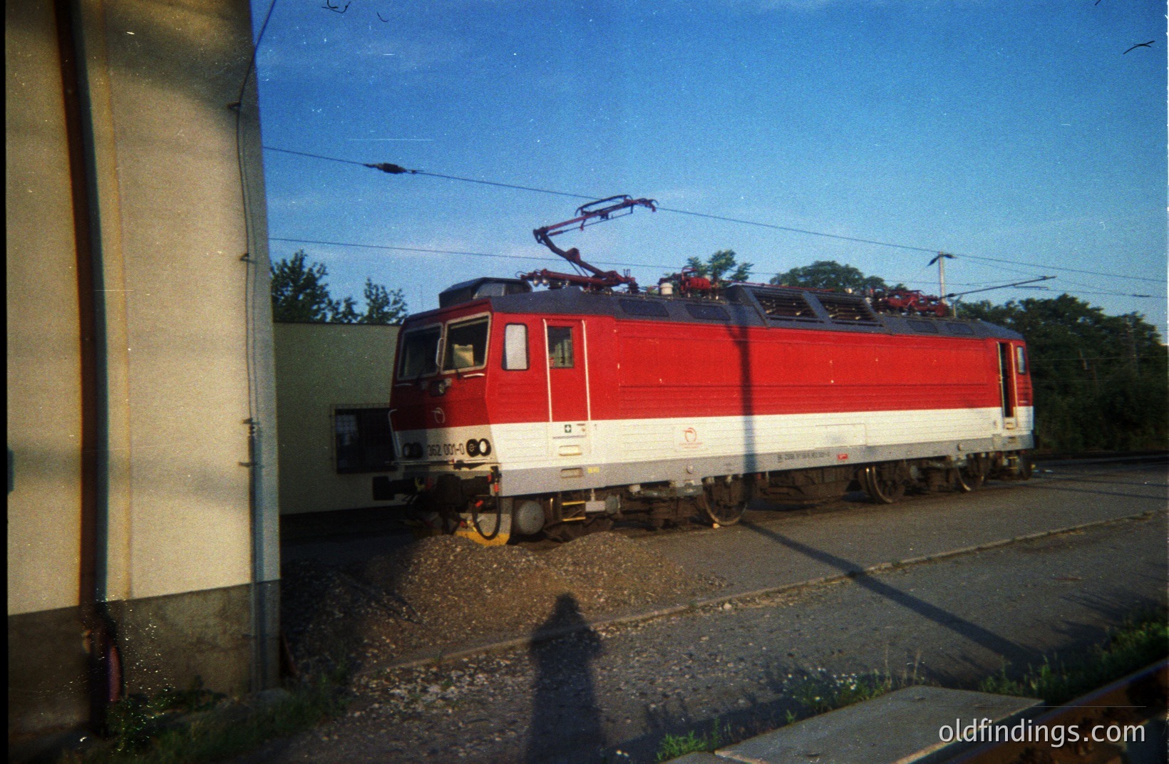 Striking, brightly colored electric locomotive stands on tracks with overhead power lines visible. Red and white livery indicates likely European origin, potentially 1970s-80s. Appears well-maintained. Likely industrial or rail yard setting.