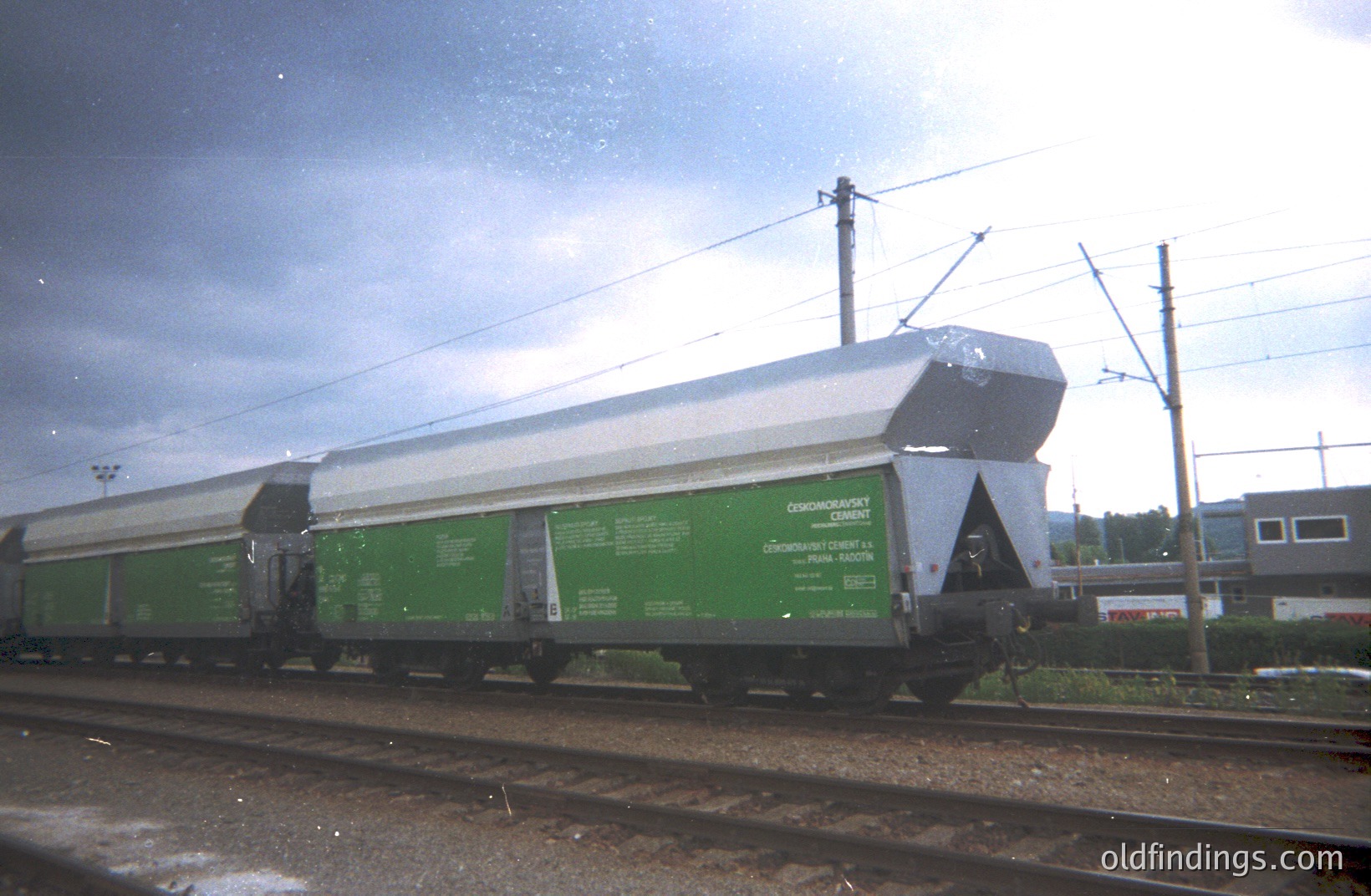 A long, green freight rail car carries stacked slabs of what appears to be precast concrete, likely for construction. The car's markings indicate its origin or destination. Overhead power lines run alongside the tracks. A building is visible in the background. Likely mid-20th century.