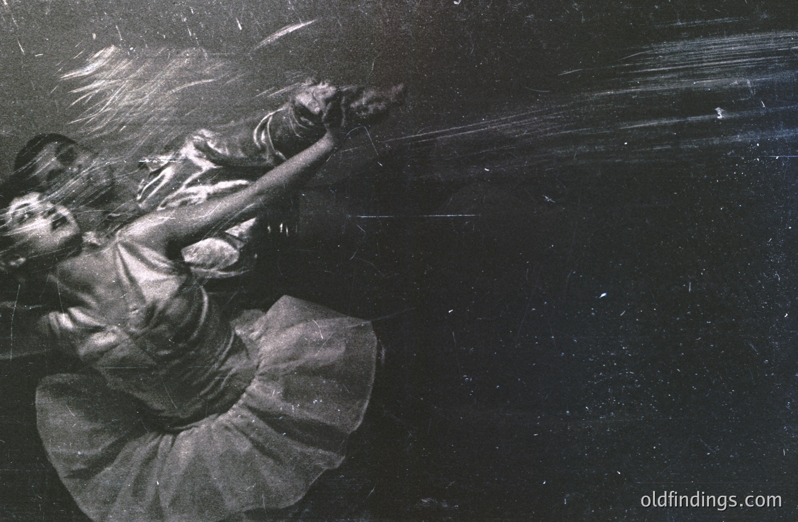 A dynamic, blurred monochrome photograph captures a dancer in mid-motion, likely a pirouette. The dancer wears a tutu and ballet flats. Strong lighting creates dramatic shadows and highlights, emphasizing movement and form. The image exhibits significant grain and scratches, suggesting age and potential archival origin. Likely 1960s-1970s ballet photography.