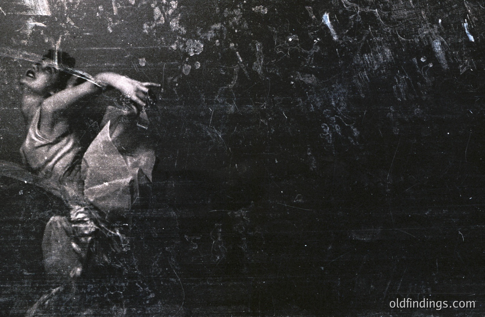 Striking, dramatic dance portrait of a figure in mid-motion, likely female, with flowing fabric emphasizing movement. Grainy monochrome highlights expressive pose and intensity. Heavily distressed and scratched surface suggests age or intentional artistic effect. Potential 1970s aesthetic.