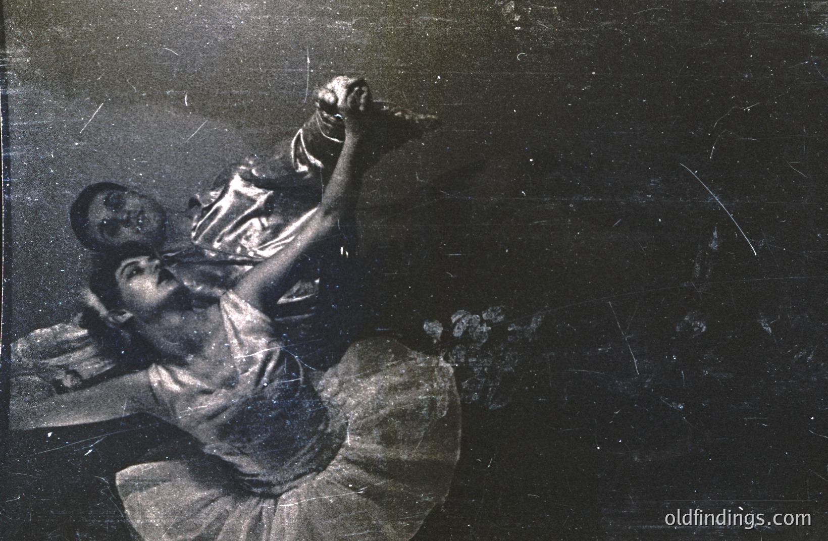 A dancer in a flowing gown is captured mid-performance, seemingly lifted by a partner who is mostly obscured. The image exhibits visible grain and wear, suggesting age and potential archival value. Likely a studio or theatrical setting.