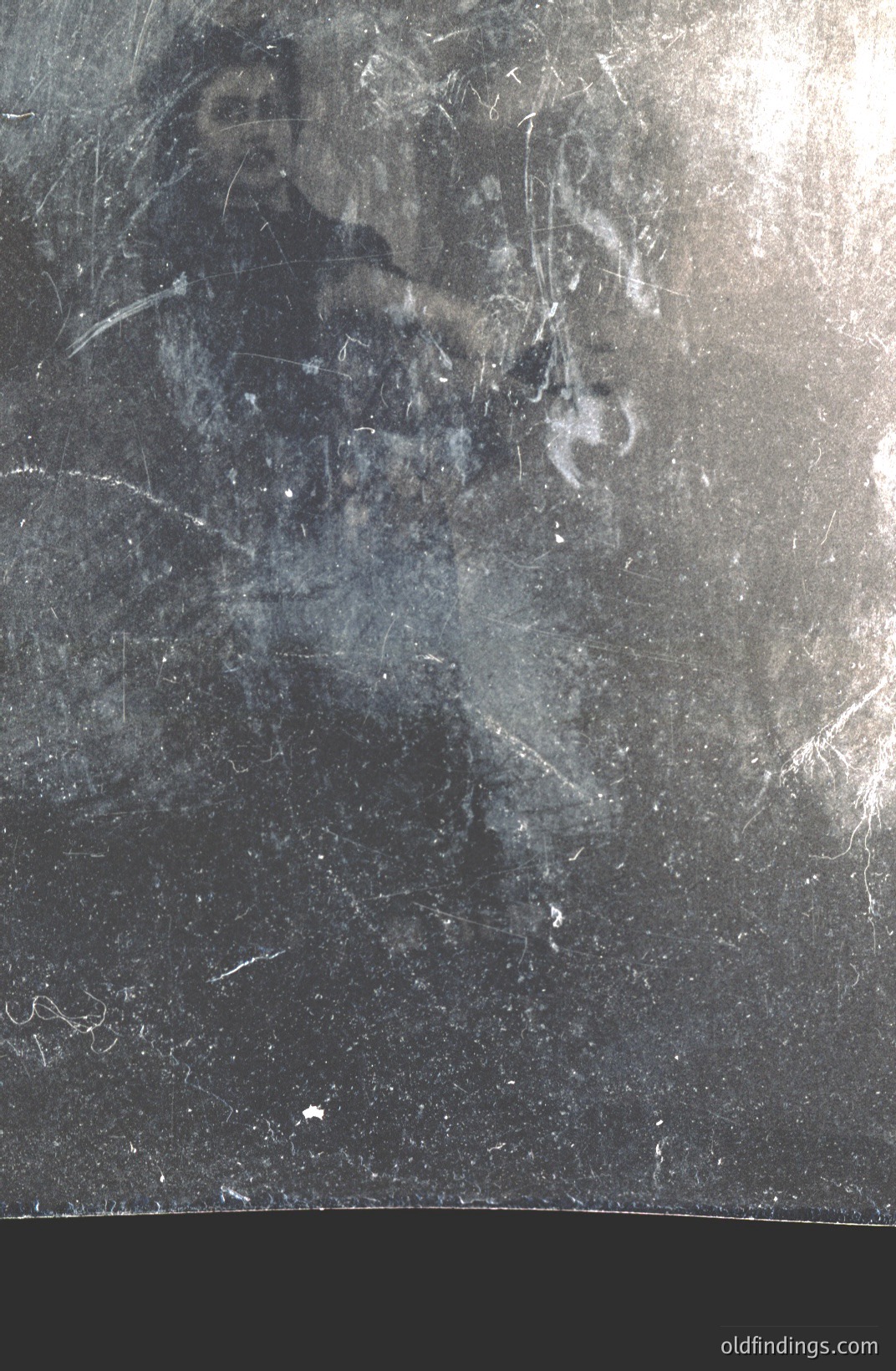 This is a close-up photograph of a weathered, dark-colored surface, likely aged wood or painted metal. Visible are numerous scratches, cracks, and areas of wear revealing underlying textures. A faint, ghostly impression resembling a human face is discernible amidst the damage. Likely a deteriorated or damaged archival print.