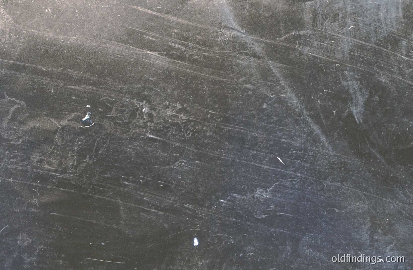 Dust and scratches across a dark grey surface dominate this image. The texture suggests aged photographic film or an oxidized metal plate. Streaks and marks indicate handling or environmental exposure. Potential use for texture overlays or vintage design elements.