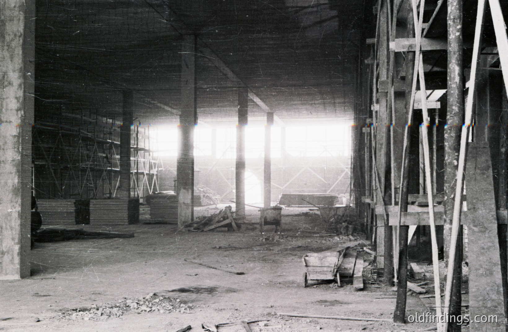 Striking black and white image shows a vast, unfinished industrial space. Concrete pillars support a high ceiling, with scaffolding visible in the distance. Construction debris, including a wheelbarrow and lumber, are scattered across the floor. Likely a mid-20th century construction site.