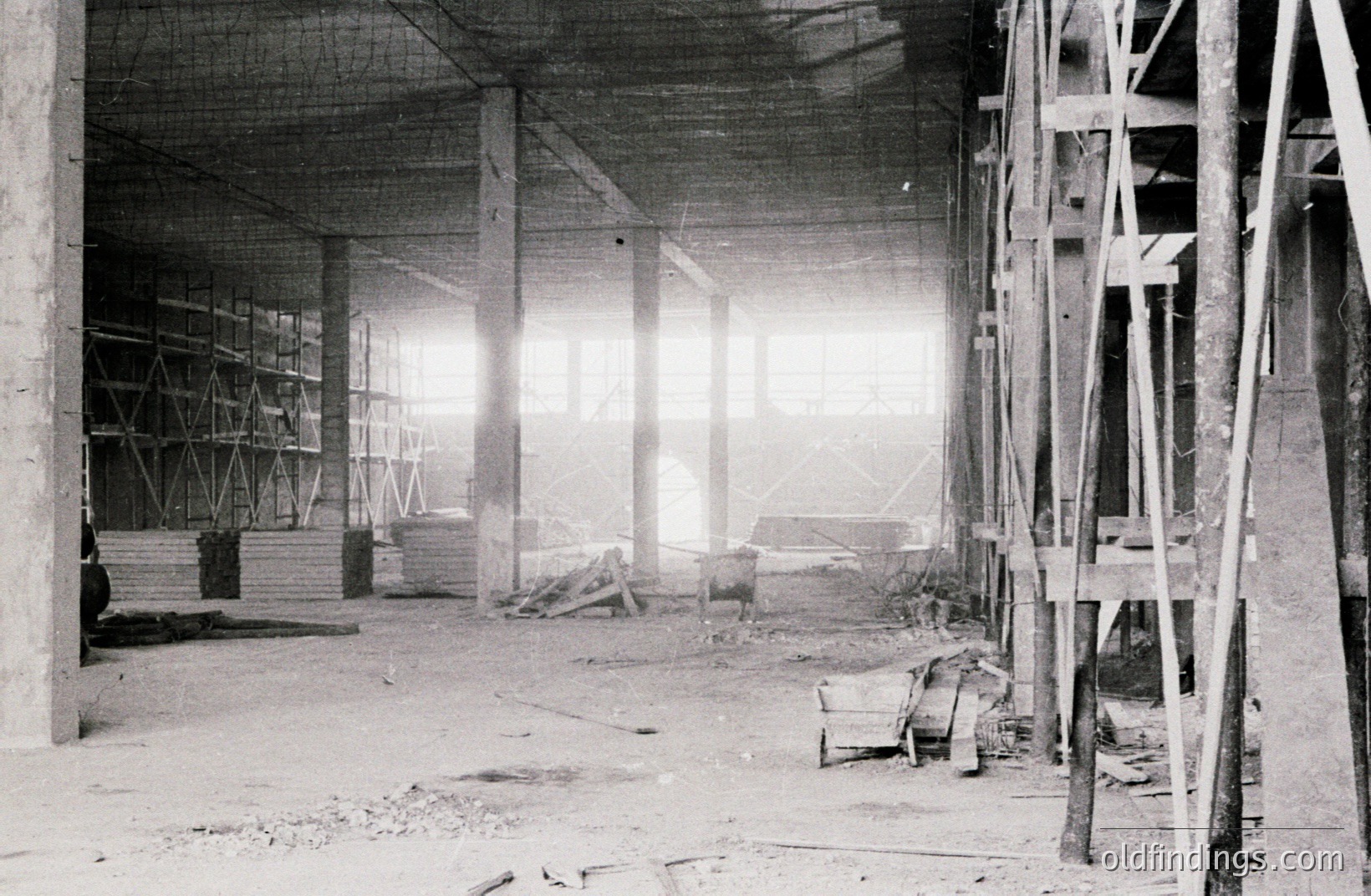 Striking black and white photo depicts a construction site interior. Concrete pillars and scaffolding dominate the view, hinting at an expansive, unfinished structure. Visible lumber and a discarded chair suggest active, yet paused, work. Likely mid-20th century, reflecting post-war construction boom.