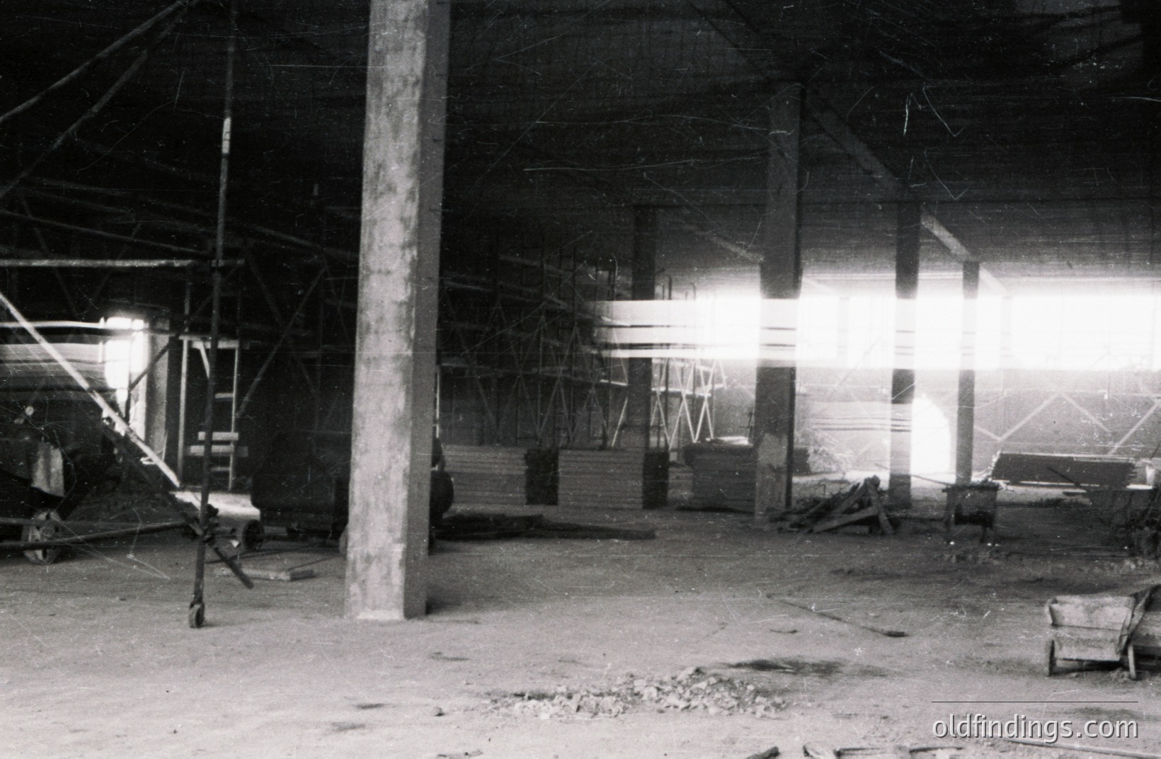 An interior view depicts a construction site, likely unfinished. Concrete pillars support a timber-framed roof structure. Lumber, scaffolding, and a wheelbarrow are visible. Dust and webbings cover surfaces. Appears to be a 1950s-1970s photograph. Suitable for historical research or design references.