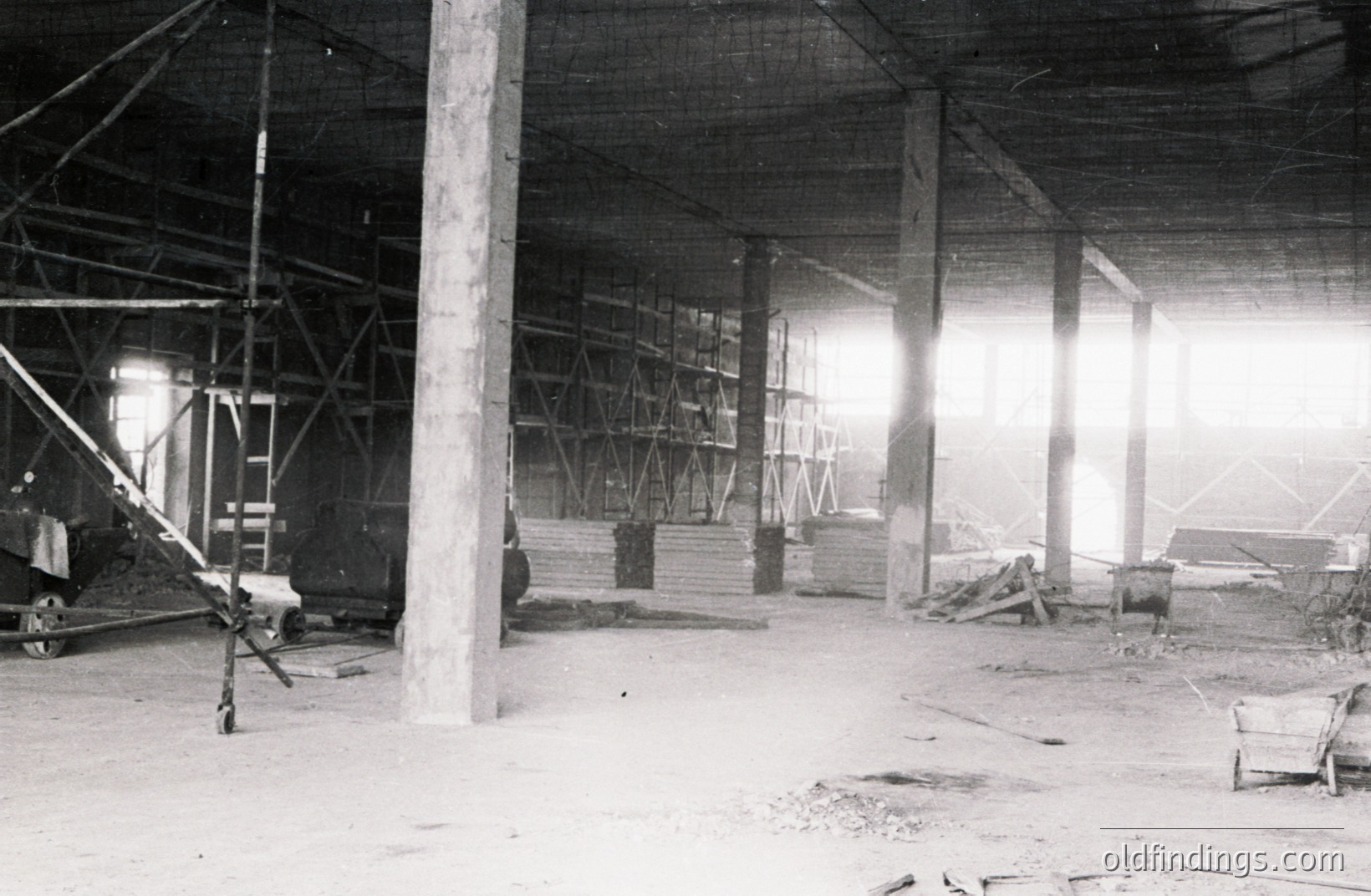 An industrial interior shows stacked lumber, supporting pillars, and a complex system of scaffolding. Appears to be an unfinished warehouse or factory construction site. Light streams in from an unseen opening. Likely mid-20th century. Potential for design/architecture reference.