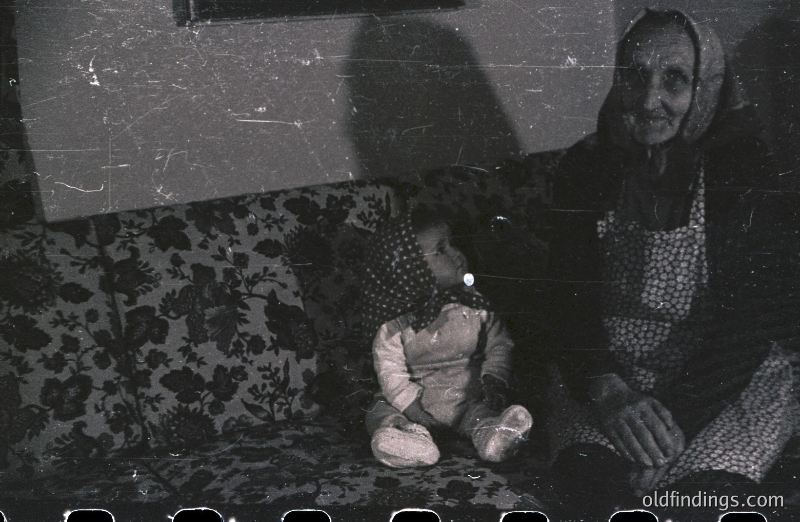 An elderly woman sits with a child on a floral-patterned couch. The scene is dramatically lit, creating strong shadows that obscure details. Possibly a family portrait, potentially from the mid-20th century. Grainy film stock and damage suggest archival value.