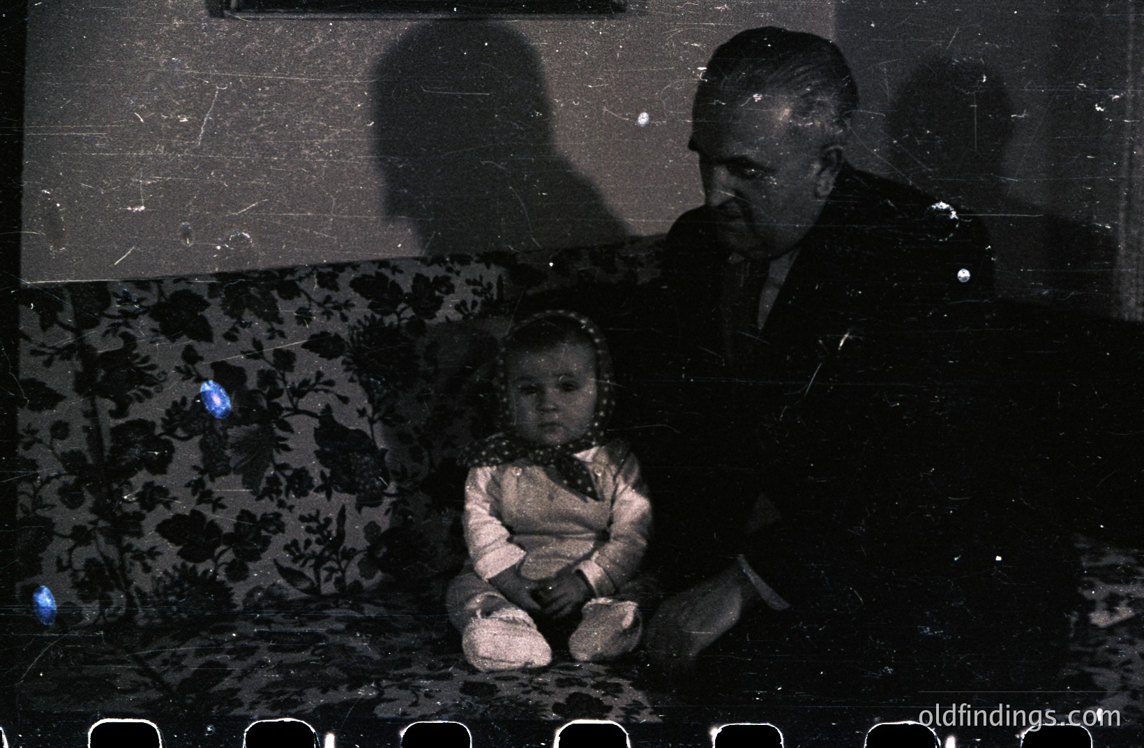 A seated man, in a suit, leans toward a young child wearing a patterned dress and a bow. They are positioned on a floral-patterned sofa, a shadow looms behind them. Likely a family portrait from the mid-20th century. The image exhibits film grain and dust.