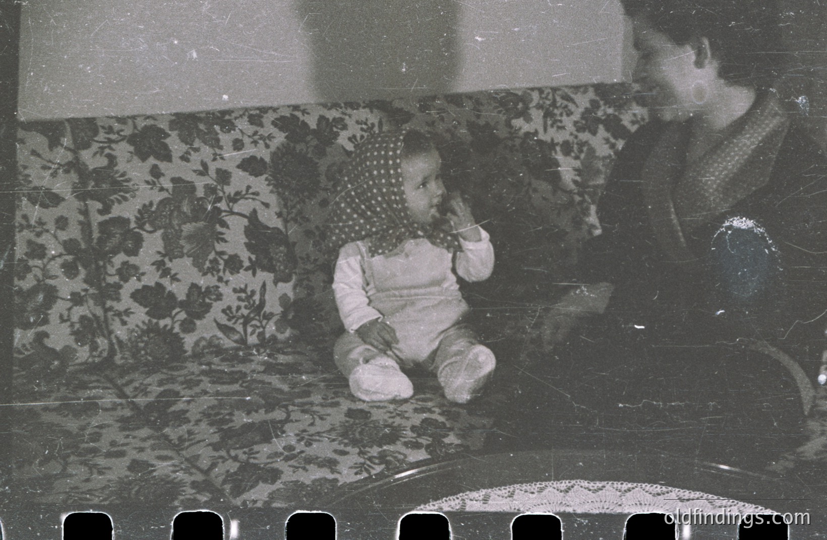 A baby wearing a patterned bonnet sits on a floral-upholstered sofa, gesturing towards a woman seated nearby. Likely a candid family snapshot, showcasing everyday domestic life. The photographic style and attire suggest a 1960s or 1970s origin. Image quality exhibits age-related degradation.
