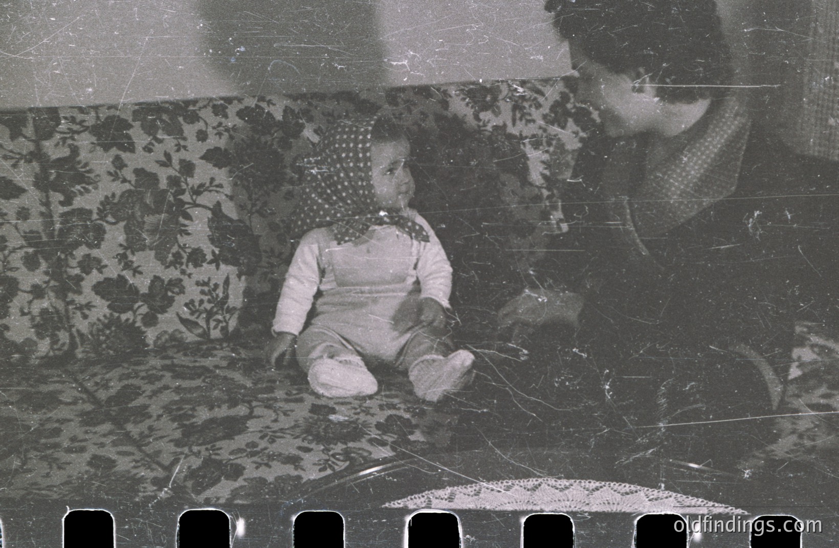 A seated toddler, clad in a romper and headscarf, is held by an adult, likely a parent. Detailed floral upholstery is visible on the sofa behind. The photograph's grainy texture suggests a vintage film negative, potentially from the mid-20th century. A personal family portrait, capturing a moment of intimacy.