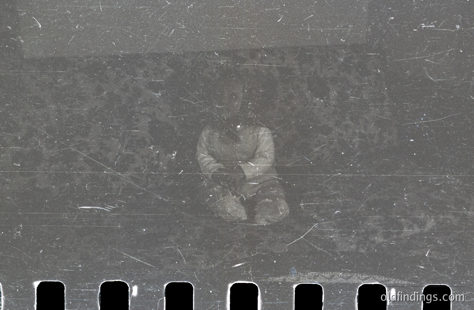 A blurred figure appears on a strip of 35mm film, showcasing noticeable dust and scratches characteristic of aged photographic material. The individual is seated, suggesting a portrait or candid capture. Film's sprocket holes are visible along the edge. Likely a reference image for archival purposes or artistic design.