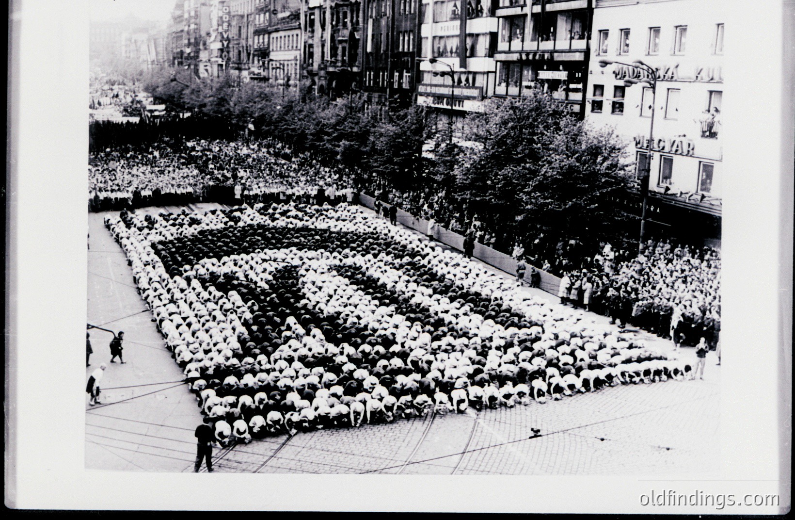 Crowd forms a large, grid-like pattern on a paved plaza. Tall buildings frame the scene, with a signage visible at the upper right. The formation appears organized, possibly for a public display or event. Likely mid-20th century, archival photograph. High commercial value for historical, design reference.