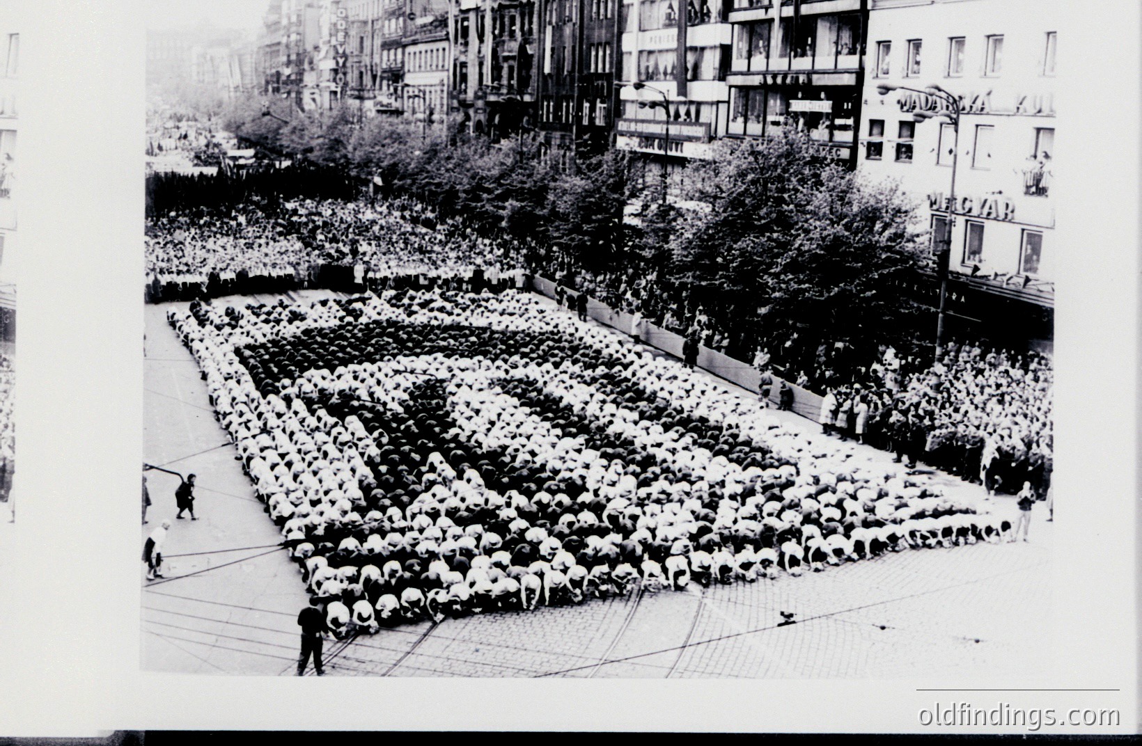 A vast, meticulously arranged group of people forms a stylized human image within a city square. Buildings flank the scene, with people lining the elevated walkways. This appears to be a planned public event, possibly a performance art piece or a promotional display. Appears to be mid-20th century.