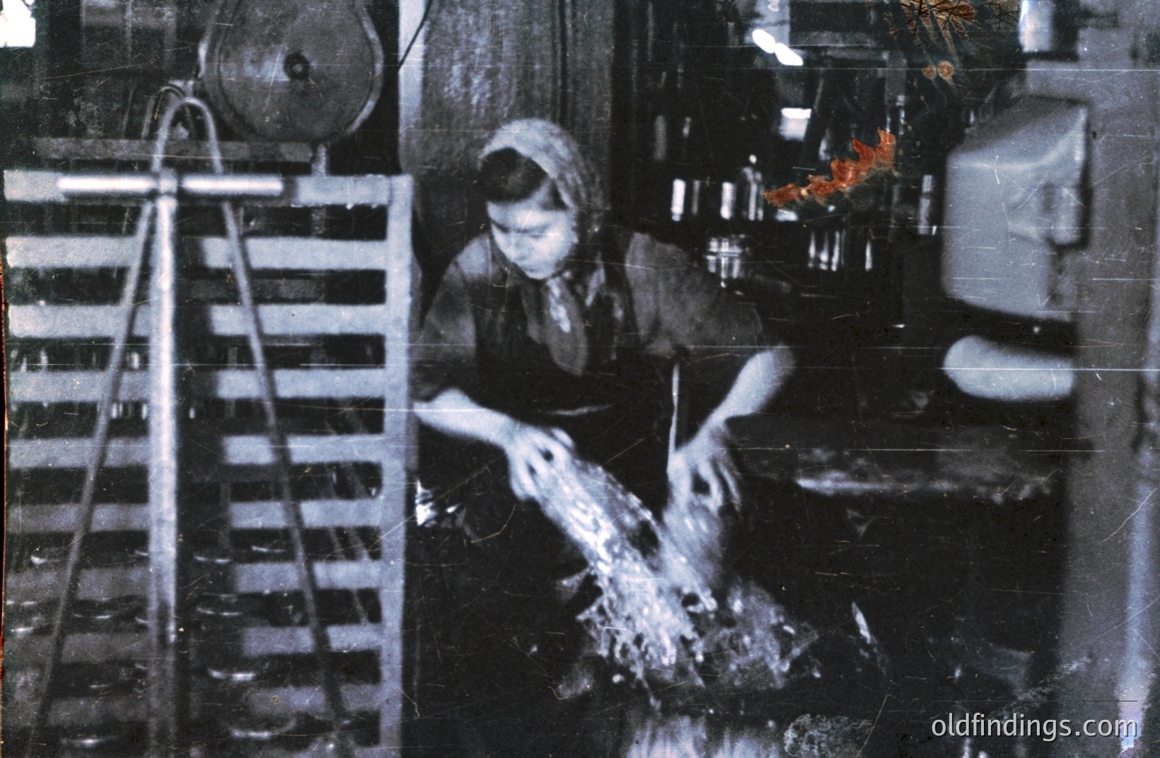 A woman in traditional dress appears to be processing a substance in a factory setting. Metal machinery, including a hopper and pipes, dominates the background. Likely from the mid-20th century, this photo captures industrial labor and process. The image exhibits signs of aging with visible scratches.