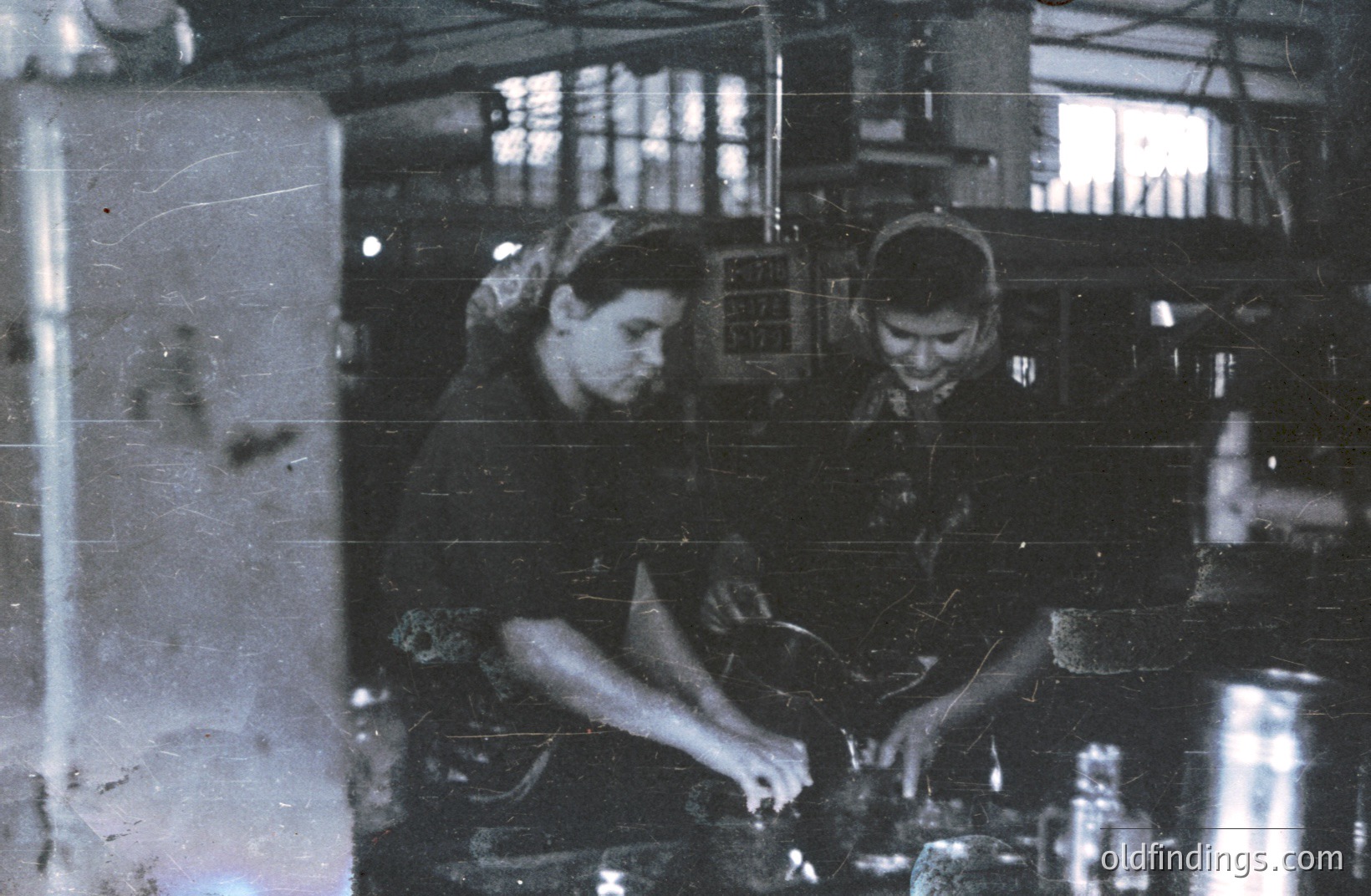 Two young factory workers appear focused on their tasks in a darkened industrial setting. Evidence suggests metal fabrication or a similar manufacturing process, indicated by machinery. The image’s grainy quality suggests it’s a vintage print, possibly from the 1970s or 80s. Possible use for historical labor studies.