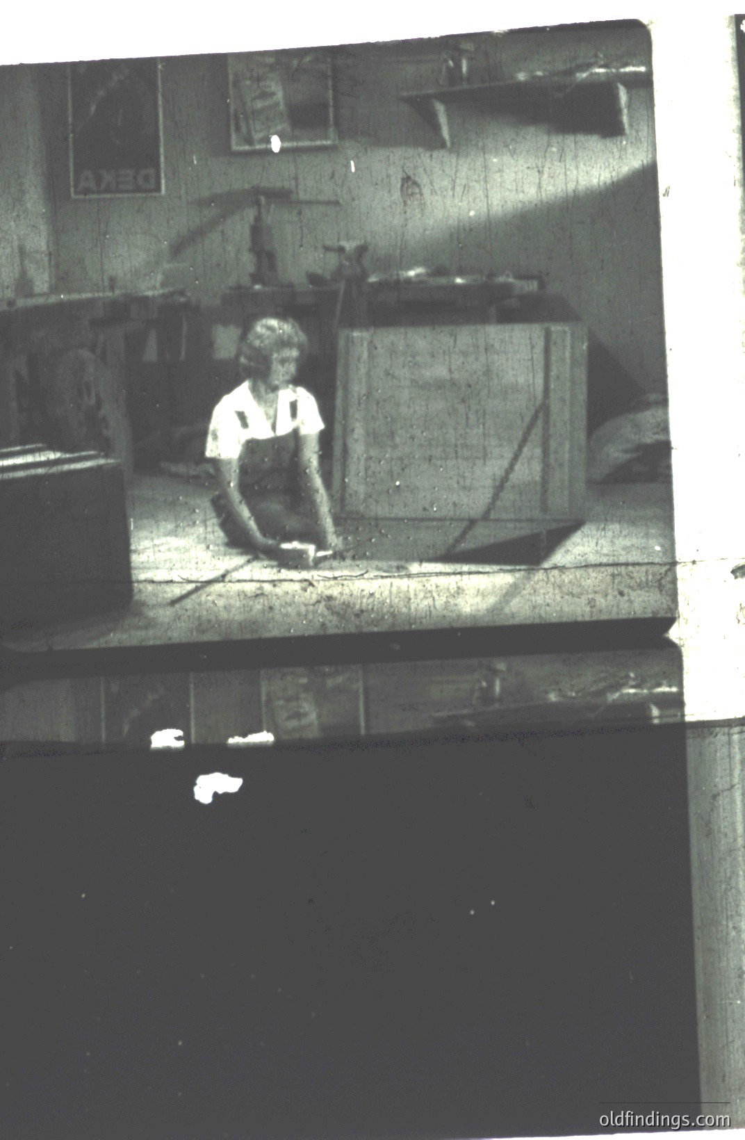A figure in work overalls kneels, seemingly examining a floor surface within an interior space with visible structural beams and stacked boxes. Likely a workshop or storage area. Possible 1960s-1970s era, judging by photographic style and clothing. Commercial value: potential for design reference, industrial aesthetic.