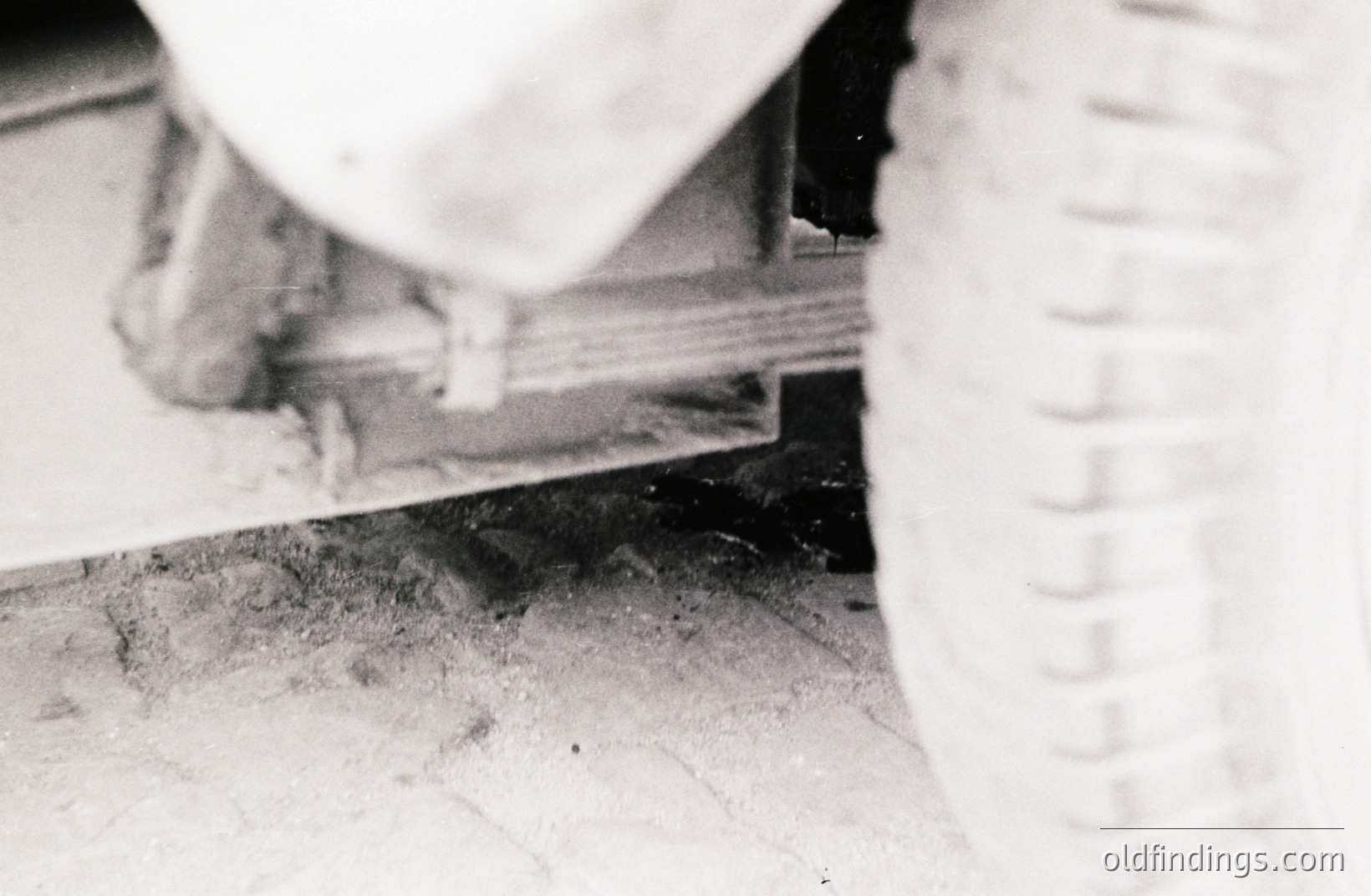 Low-angle view capturing part of a vintage vehicle's tire and chassis. The tire shows detailed tread, indicative of a utility or off-road vehicle. A brick or stone floor surface is visible, creating texture and shadow play. Likely a 1950s-1970s image.