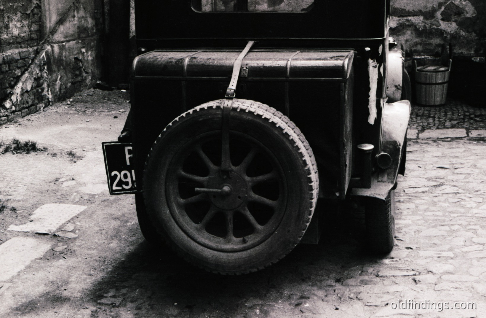 Vintage car rear view with visible license plate "F 29". Features include a wire-spoke wheel, period-appropriate tire, and rudimentary mudflap. Set against a weathered stone wall & cobbled ground. Likely a 1920s or 1930s vehicle. Ideal for design or automotive history reference.