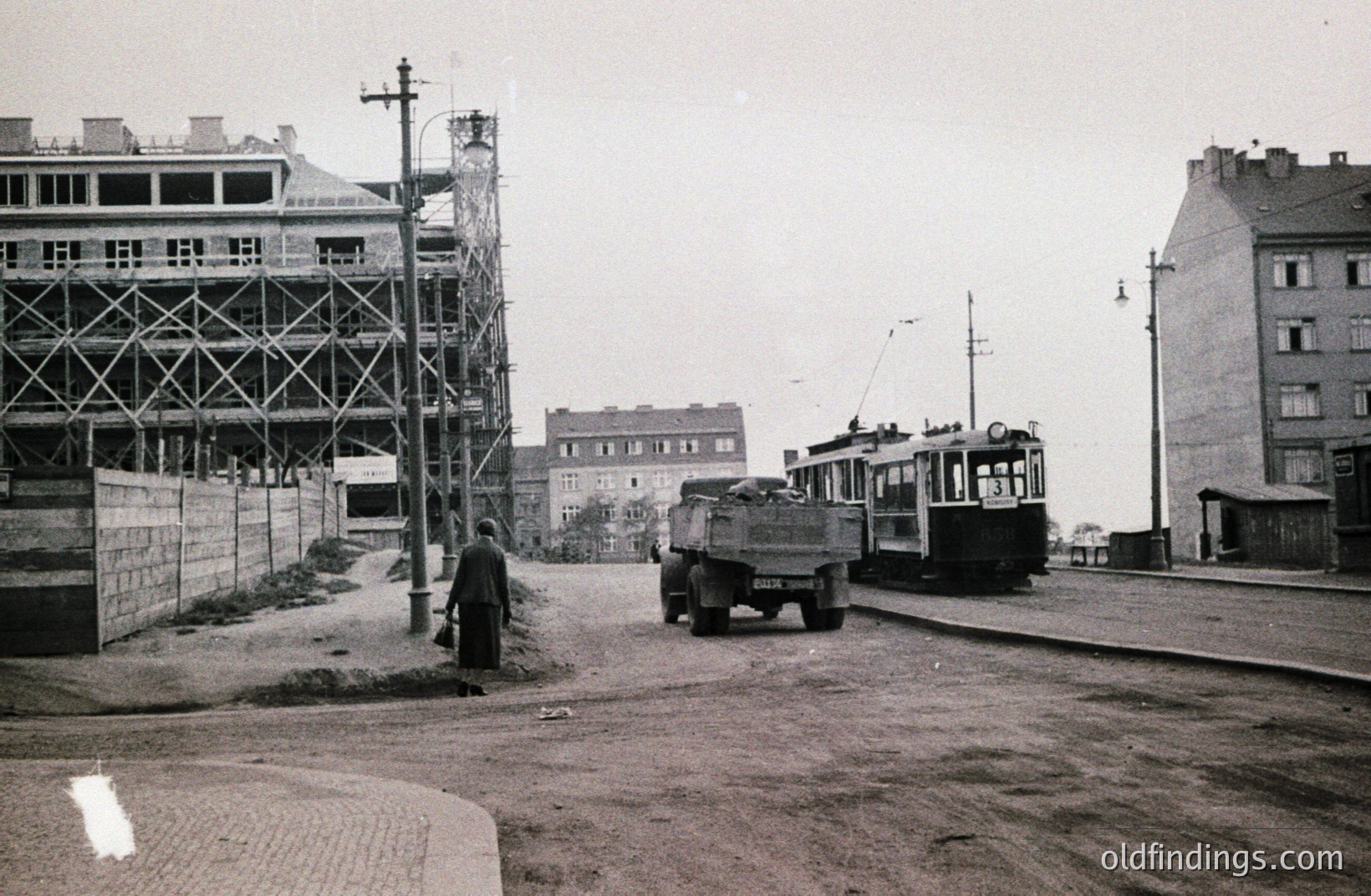 Construction site & tram meet on a street. A man walks past a truck, with an older-style tram in the background. Building scaffolding dominates the left side of the image. Likely post-war reconstruction, possibly 1950s-1960s Eastern Europe.