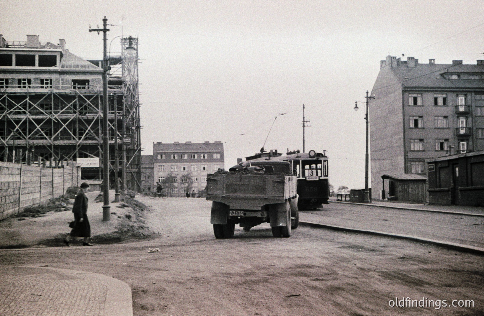 Construction site visible as a multi-story building under scaffolding dominates the view. A utility truck and tram are on a street lined with older residential buildings. Possibly post-war reconstruction, likely 1950s Eastern Europe. A solitary pedestrian walks along a narrow sidewalk.