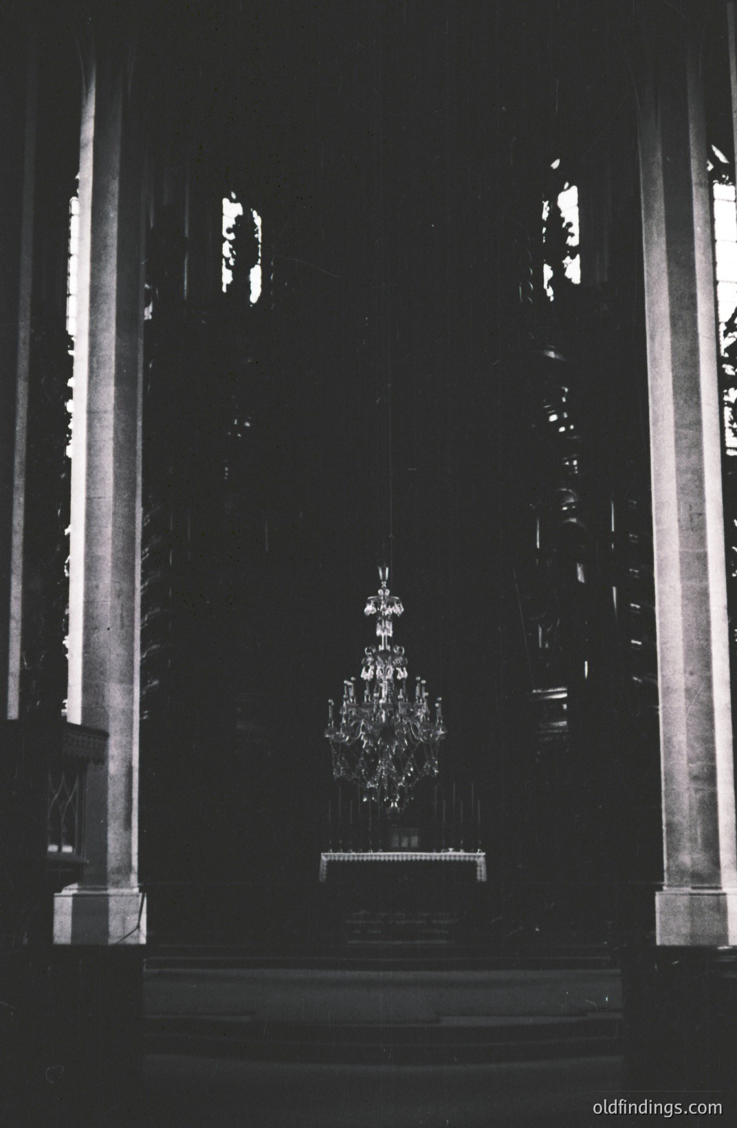 Grand, ornate chandelier hangs within a darkened, neo-classical interior. Dramatic vertical lines of columns frame the scene. The focus highlights the chandelier's reflective surfaces. Likely a church or public hall, potentially early 20th century. Excellent for design references.