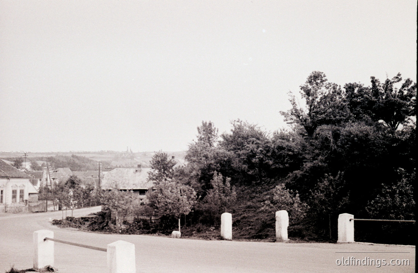A roadside scene captures a small town with traditional architecture. A stone barrier lines the foreground, framing a view of rooftops and a distant spire. Dense foliage partially obscures the landscape. Likely Eastern European, c. 1950s. Potential stock photo reference for rural landscapes.