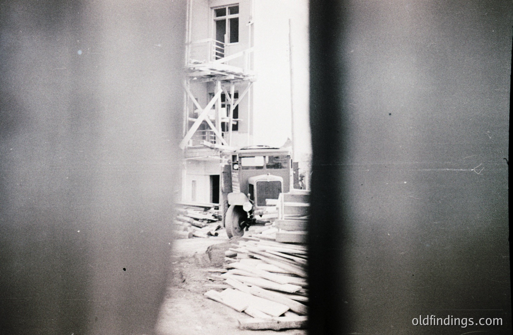 Construction scene: a truck stands amidst lumber and scaffolding before a multi-story building. Likely a mid-century commercial or residential project, possibly Eastern European based on architectural style. Visible wear and dust on the print suggest age. Valuable for architectural history and construction industry references.