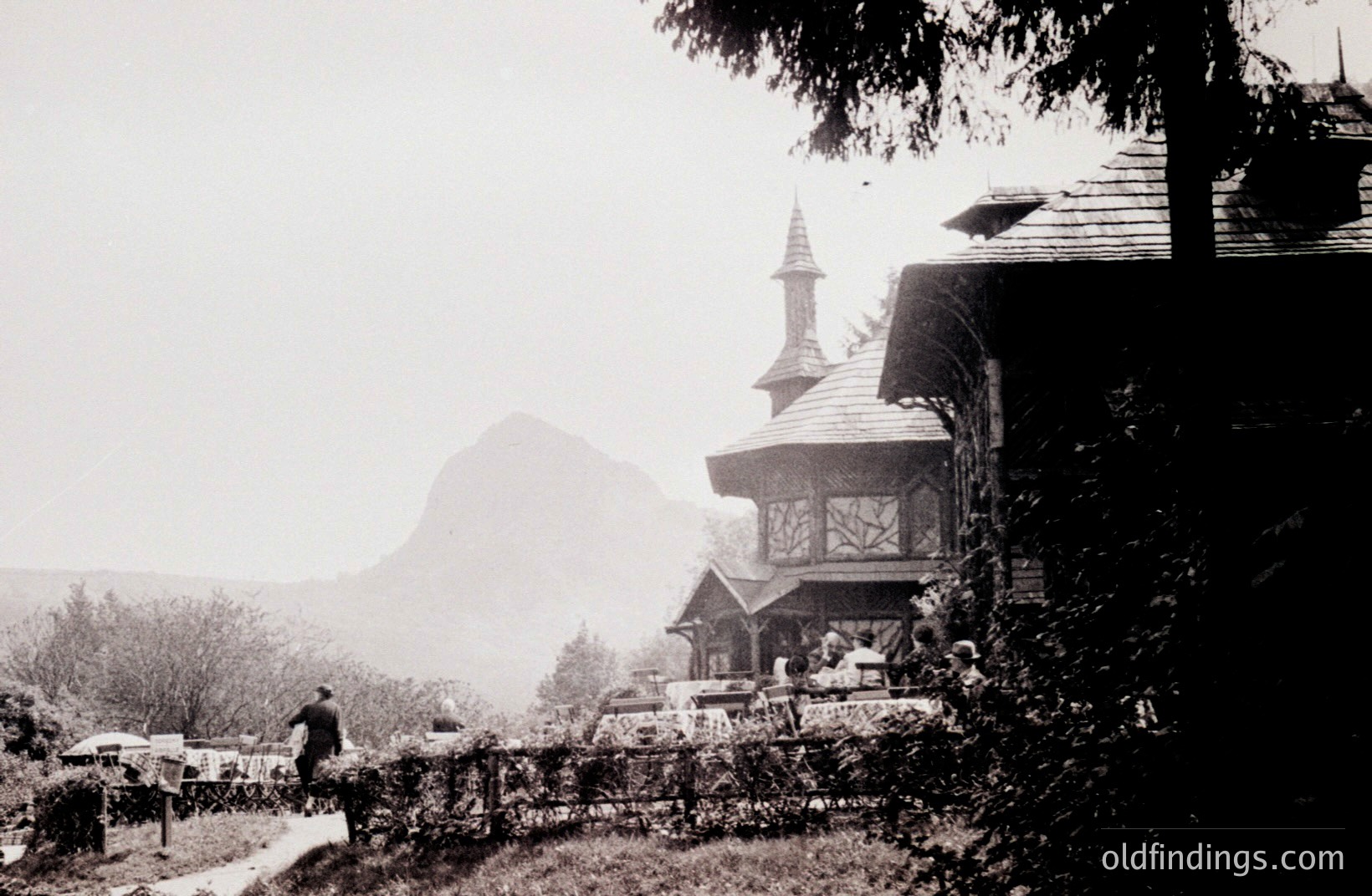Classic architectural scene: ornate wooden pavilion with peaked roof & detailed spire overlooks a landscape dominated by a pyramidal mountain. A man walks a path beside a decorative fence. Likely a resort or park setting, 1920s-1940s.