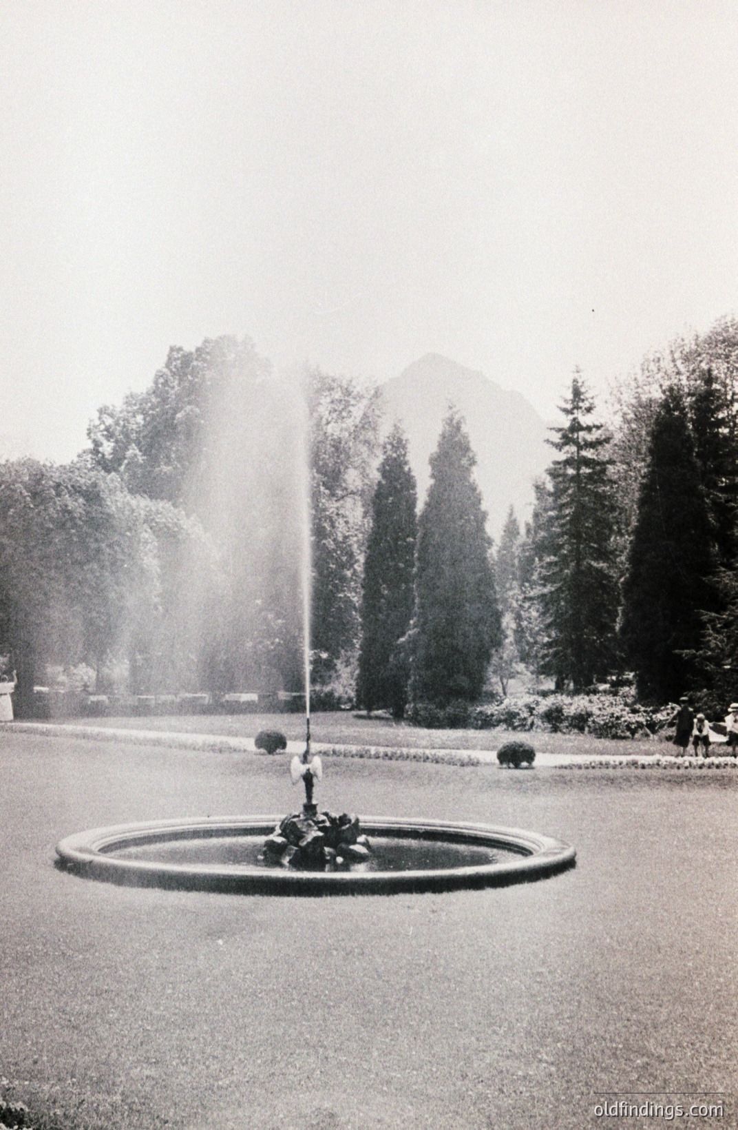 A circular fountain sprays water in a manicured park setting. Formal landscaping includes evergreen trees and a visible distant mountain range. Likely a public garden or estate. The photo’s vintage quality suggests the 1950s-1970s. Ideal for design references.