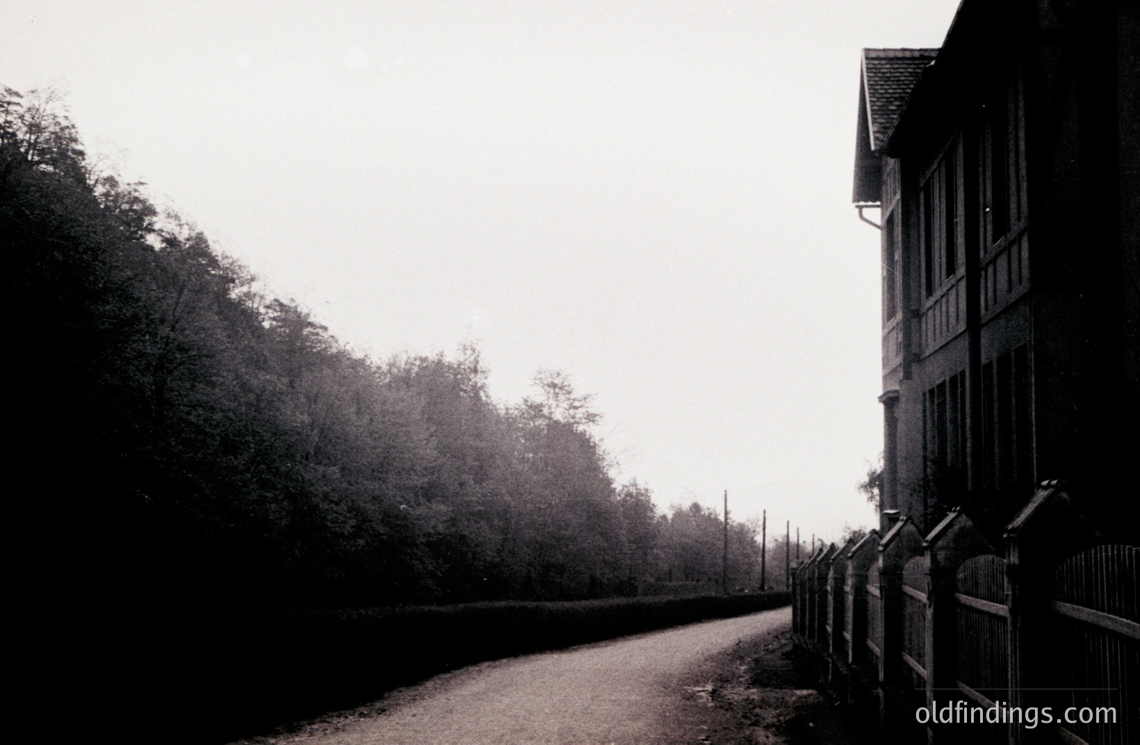 A monochrome view depicts a weathered building with dormer windows and a steeply pitched roof, bordered by a low, ornamental fence. A gravel path leads toward the structure, framed by dense trees and overcast skies. Likely late 19th/early 20th century. Could be a sanatorium or estate.