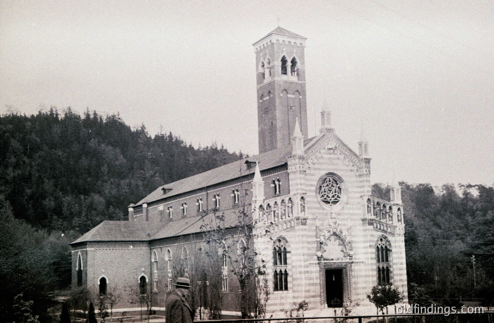 Grand Romanesque-style church with a tall, square bell tower. Detailed stone facade, arched windows, and a prominent rose window. A man in period attire stands to the side. Appears to be in a wooded, rural setting. Likely late 19th/early 20th century.
