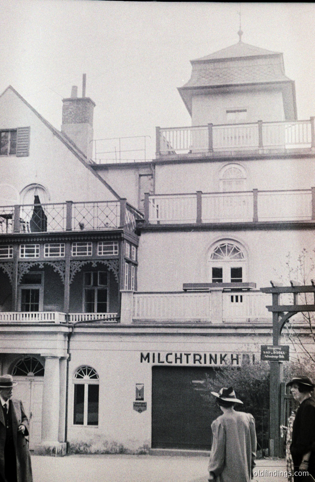 Exterior view of "Milchtrinkhalle" (milk drinking hall), a neoclassical building featuring prominent arched windows & ornate balconies. Figures in period attire are present. Likely a promotional image for a dairy or public amenity. Late 19th/early 20th century. Architectural reference.