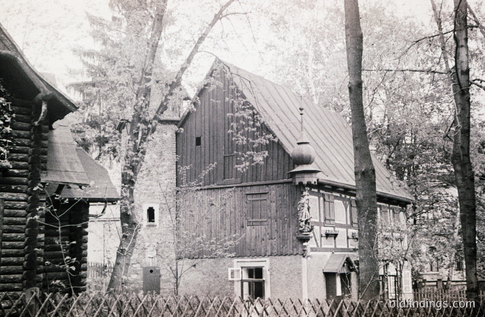 A timber-framed house with a steep, slate roof and decorative gables sits nestled amongst trees. A log cabin-style structure is visible to the left. Likely a rural or village scene, possibly Germany. Architectural details suggest a mid-20th century or earlier era. Potential stock photography reference.
