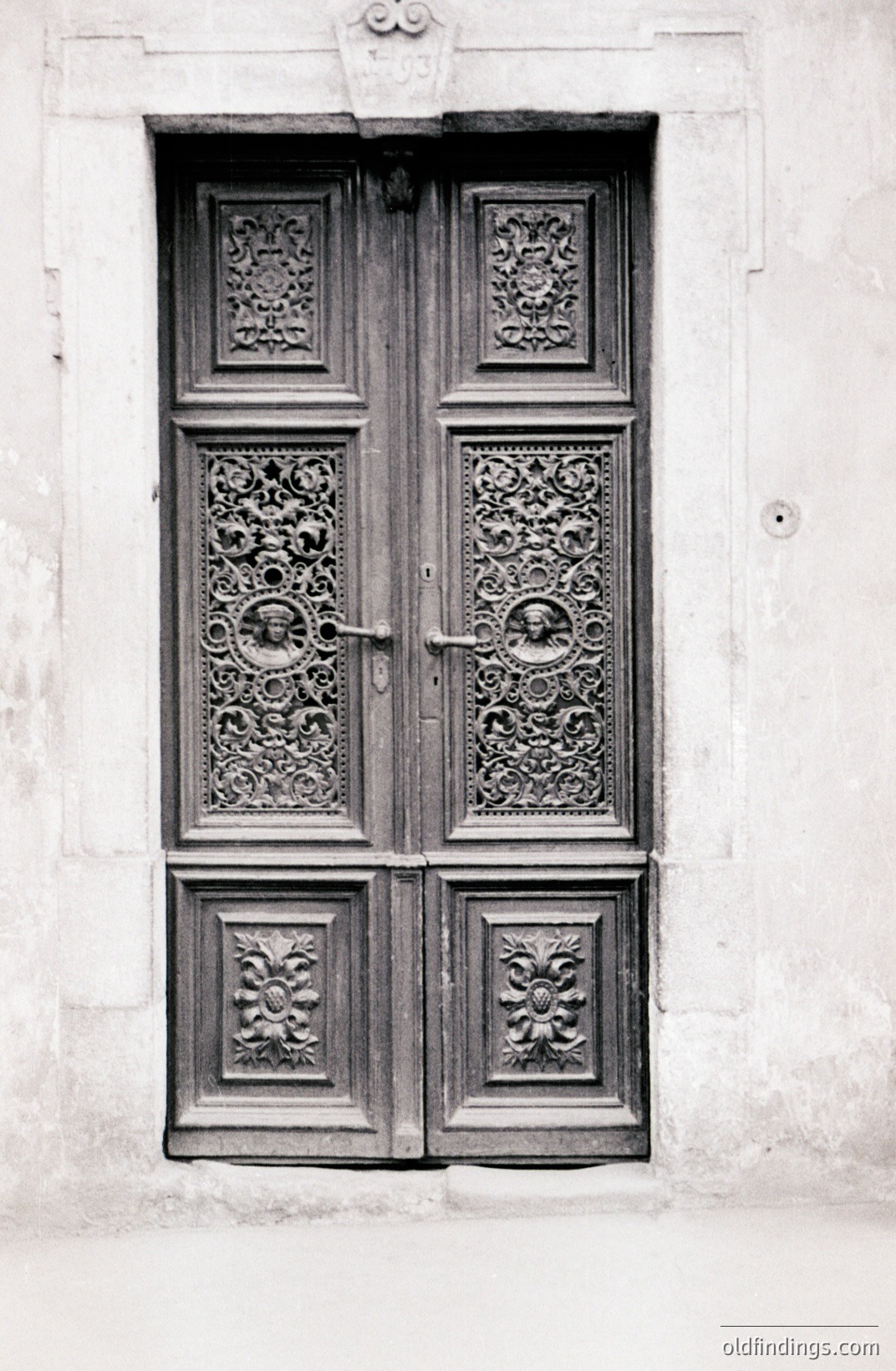 Detailed black and white image of a classical door with intricate carved floral and figurative ornamentation. Heavy, dark wood panels set within a stone frame. Likely dating to the 18th or 19th century, showcasing elaborate design details. Architectural detail suitable for design reference.