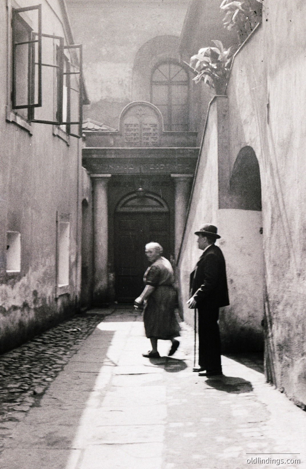 A monochrome street scene depicting an elderly couple walking toward a grand, arched doorway set within a stone facade. Paved walkway, high walls, and a visible balcony suggest a European location. Likely 1930s-1950s; architectural style hints at possible Alpine or Mediterranean influence. Commercial value as stock for historical design.