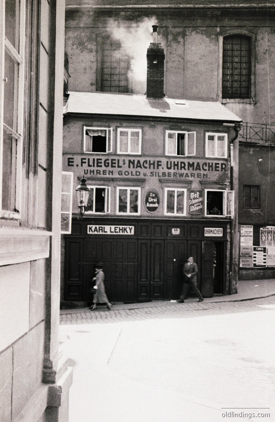 B&W street scene captures a storefront "E. Fliegel's Nachf. Uhrmacher" (watchmaker) with ornate signage & a vintage lamp. Two pedestrians pass by. The building’s architecture suggests Central Europe, possibly 1920s-1940s. Historic commercial signage offers research value.
