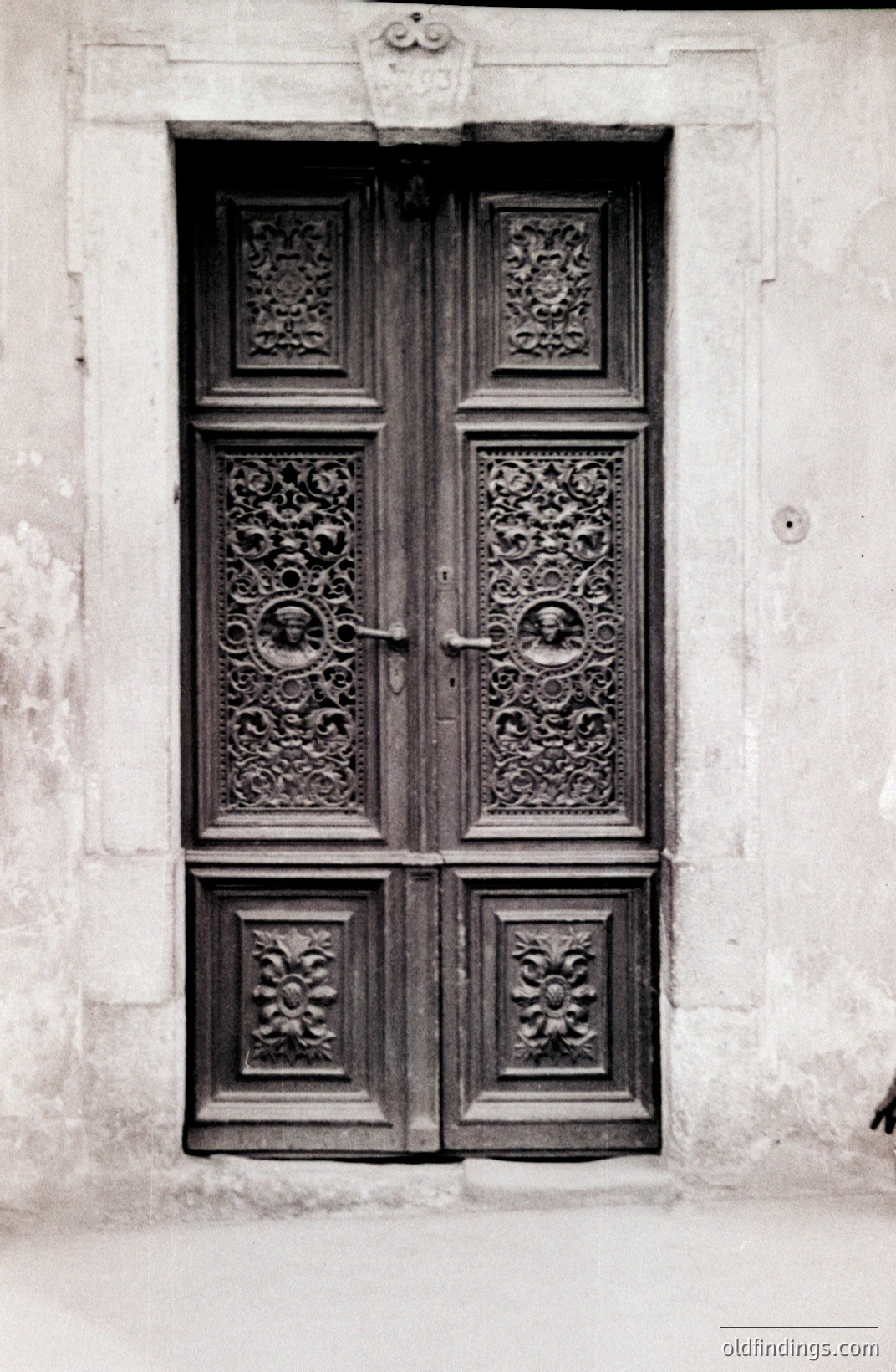 Elaborate, carved wooden doors, likely 18th or 19th century. Detailed floral and figurative relief work adorns the panels. Set within a shallow stone surround. Likely European origin; architectural style suggests Italian or French influence. A study in texture & detail.