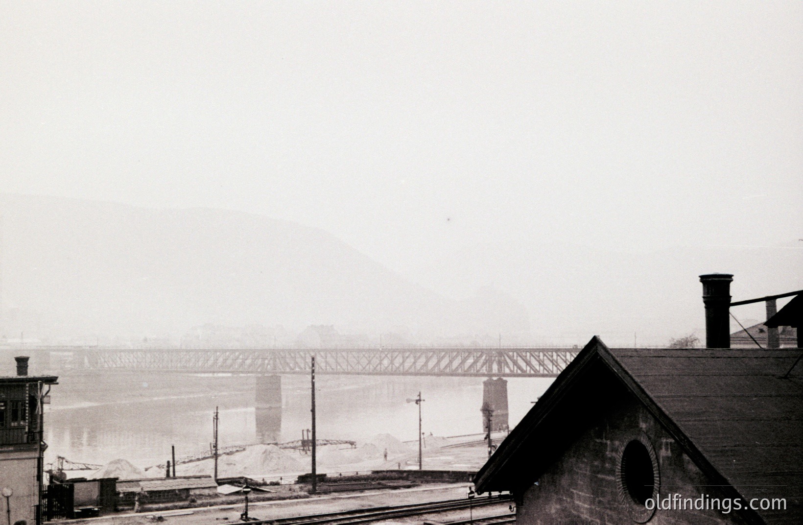 Overcast skies and fog obscure a railway bridge spanning a wide river, likely industrial in nature. A weathered building with a round window occupies the foreground. Visible infrastructure suggests a working port or industrial area. Appears to be an early 20th-century view.
