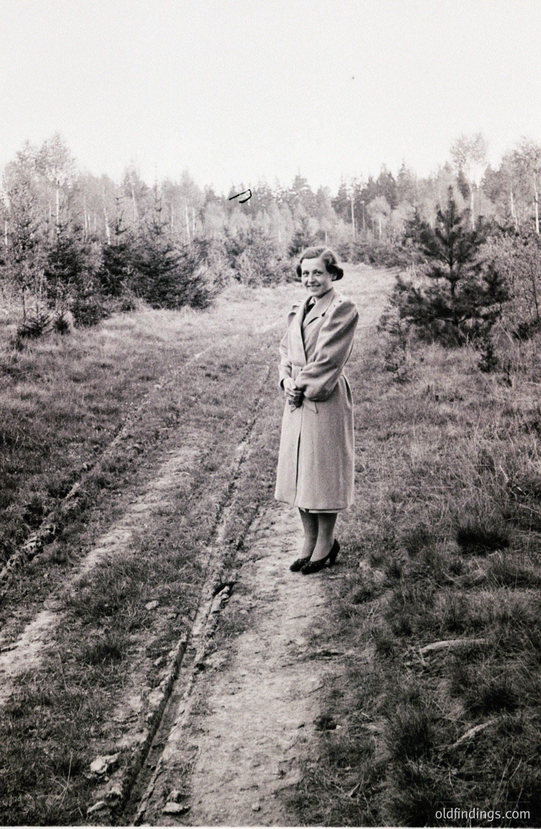A woman in a tailored coat and heels stands on a dirt track, framed by pine trees in a forested area. Likely 1950s style; the overall composition suggests a fashion or portrait study. Evokes a sense of quiet elegance amidst nature. Commercial value: vintage fashion reference.