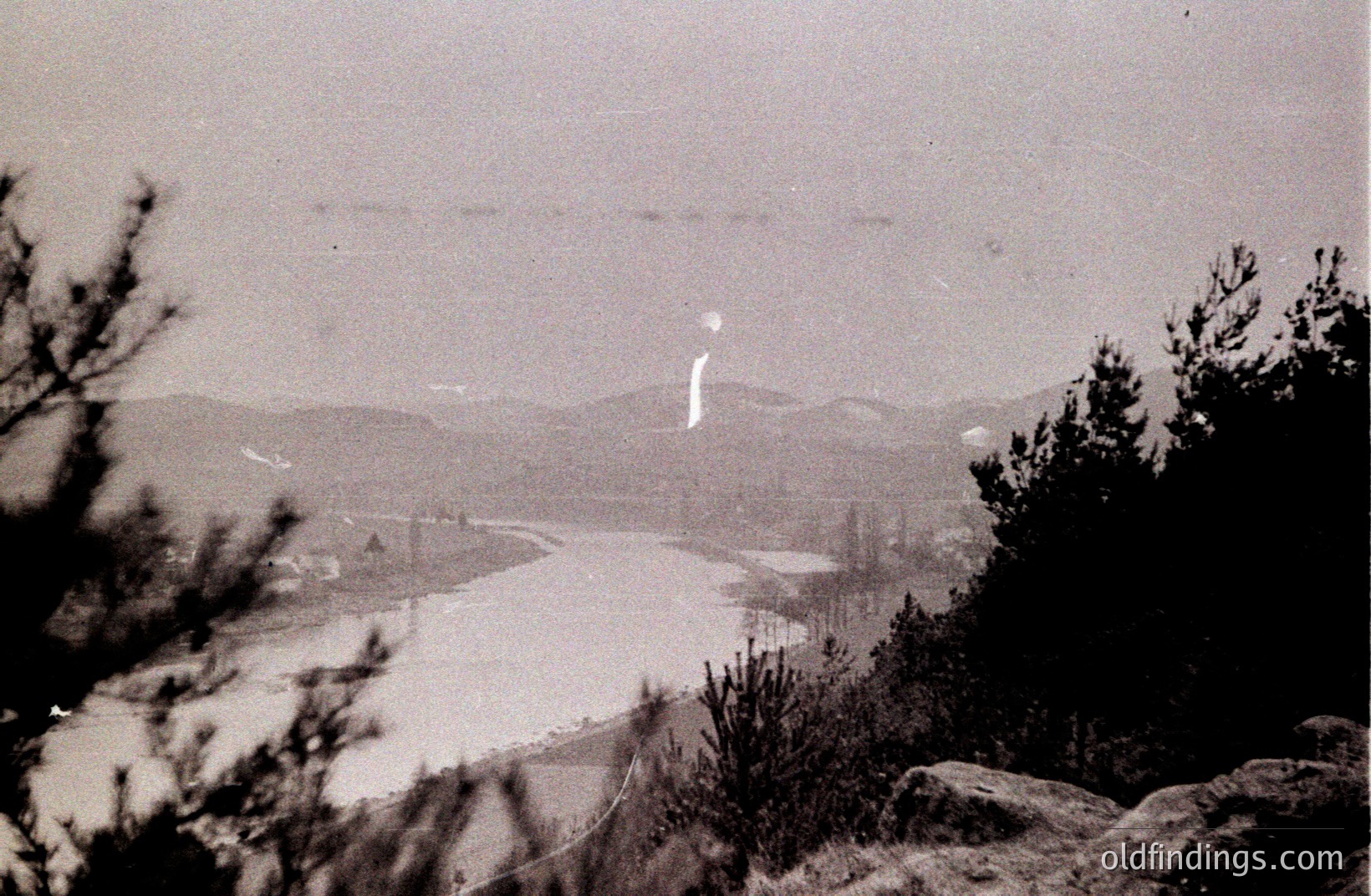 A black and white landscape photograph shows a wide, slow-moving river winding through a valley, flanked by pine forests and distant, hazy mountains. A tall, thin plume of what appears to be steam or smoke rises prominently from the riverbank. Likely from the mid-20th century.