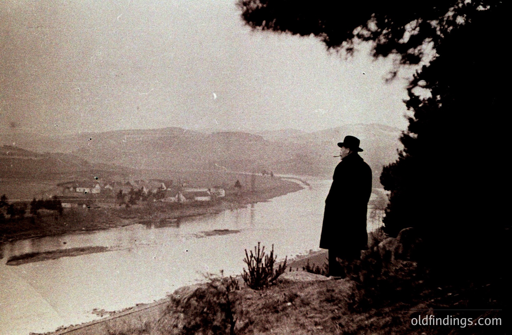 A solitary man in a dark coat and hat stands overlooking a winding river valley, likely a scenic vista. The photograph's grainy quality and clothing style suggest a possible 1920s or 1930s origin. Landscape features rolling hills, suggesting an alpine or coastal region. Composition emphasizes solitude & scale.