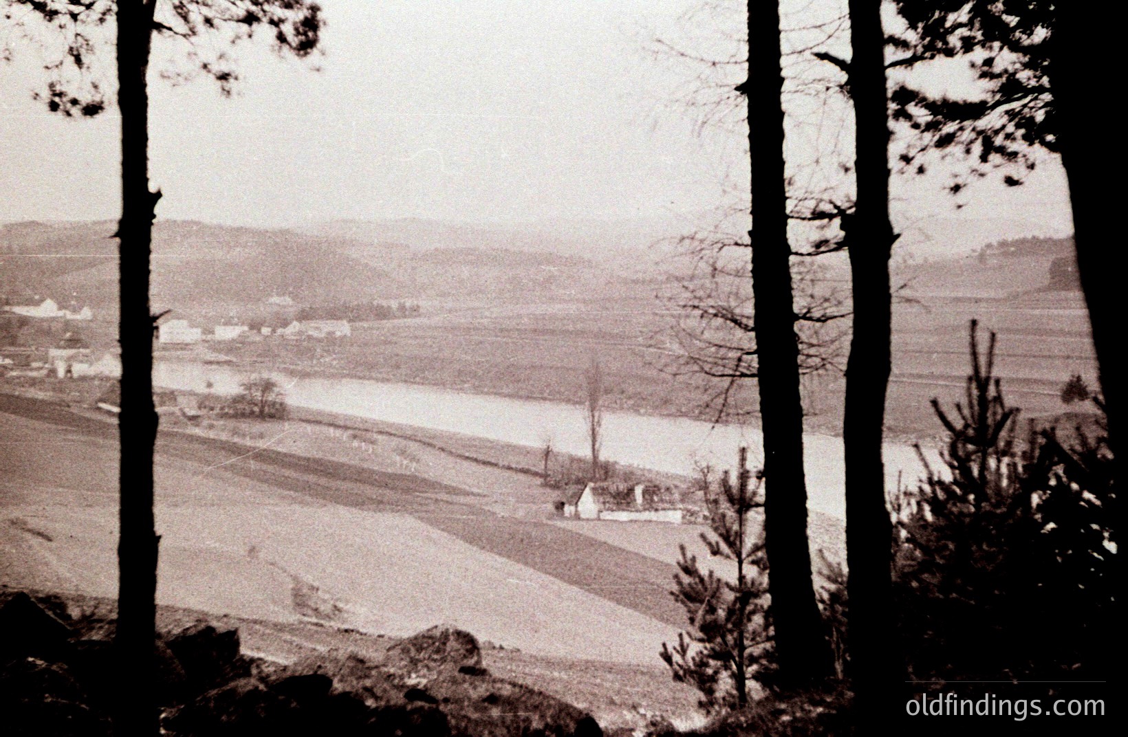 Undulating landscape with a wide, shallow river and small settlement visible through framing trees. Agricultural fields extend to the horizon, faintly patterned with rows. Likely rural scene, potentially 1950s or 60s. Suitable for design/research projects referencing pastoral scenes.