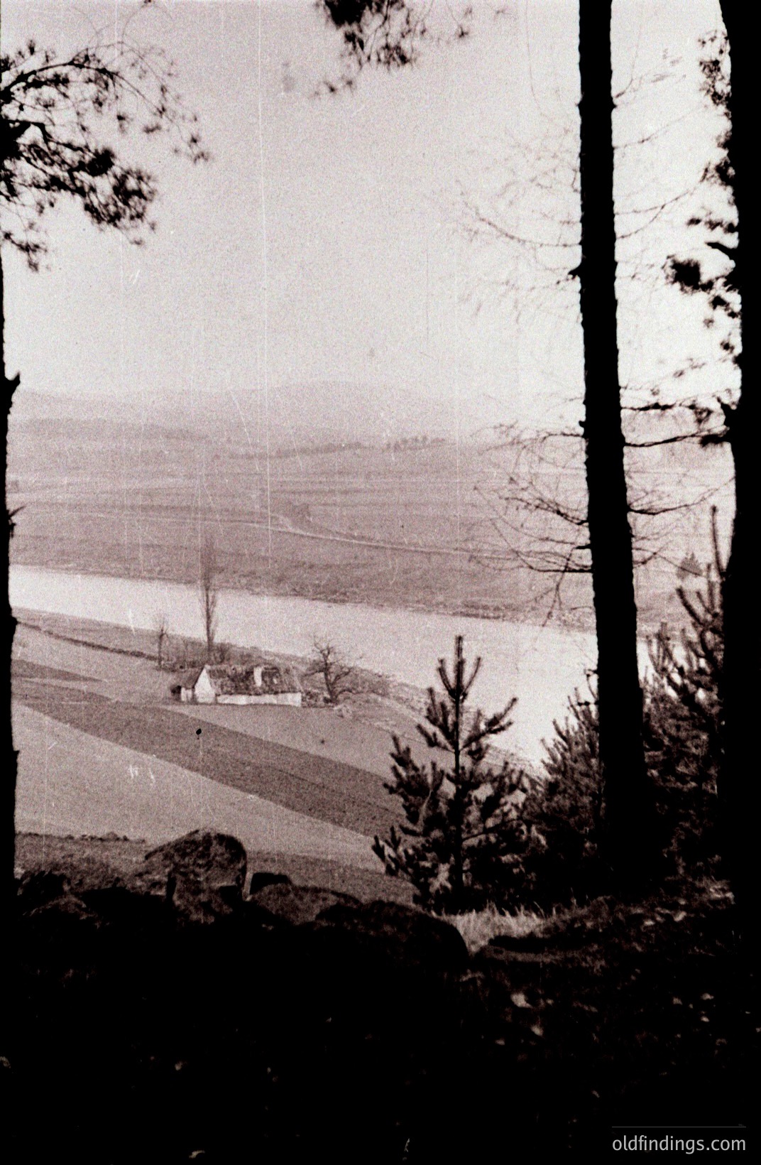 A monochrome view shows a modest building beside a wide, placid body of water, likely a lake or inlet. Gentle, terraced slopes rise in the background. Framing branches suggest an elevated vantage point. The image exhibits signs of age, including visible grain and surface wear. Likely mid-20th century.