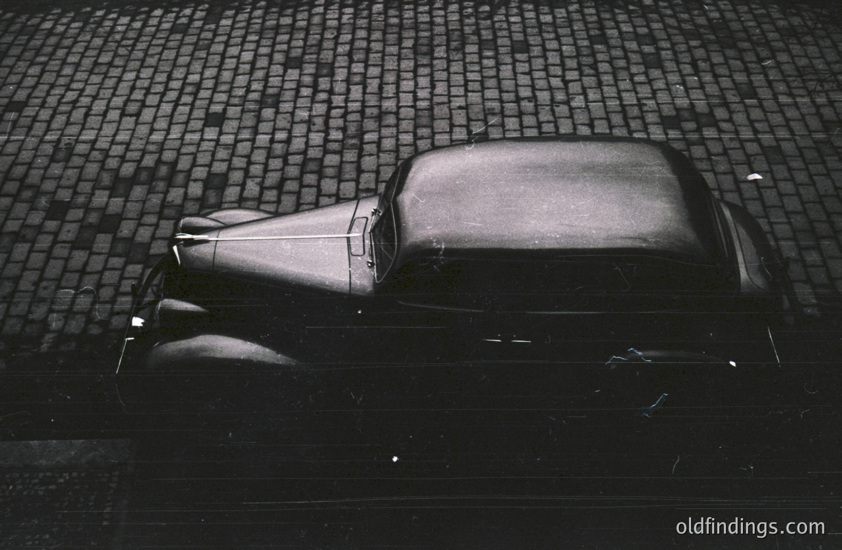Classic car, likely a 1930s model, photographed from above on a cobbled street. The vehicle's dark color and angular design are striking against the patterned pavement. High contrast highlights the car’s form. Potential stock value for automotive design or vintage aesthetics.