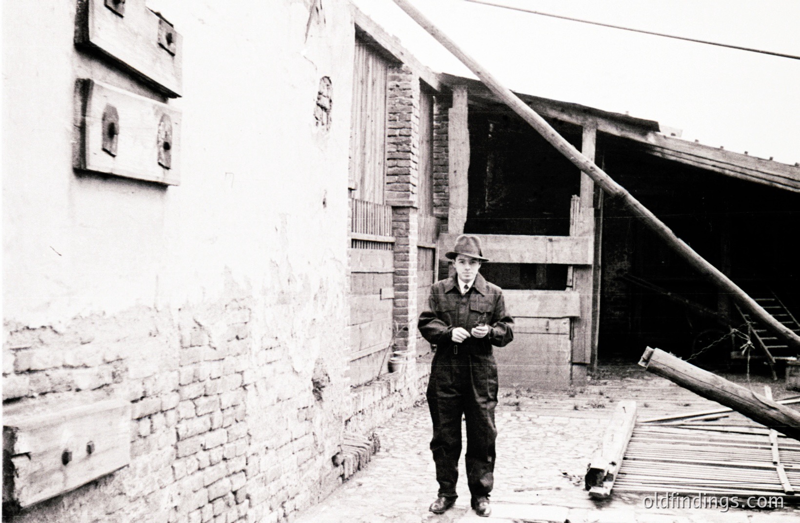 A young man in a dark suit and hat stands outside a weathered brick and timber building. The structure shows signs of age and disrepair with exposed beams and a tilted roofline. The man appears to be examining something in his hands. Likely 1930s or 40s.